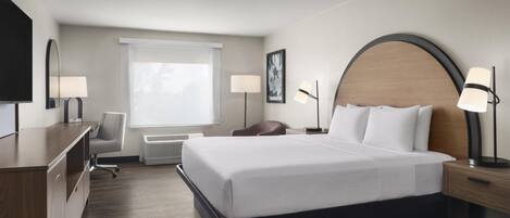 A hotel room with a large bed, bedside lamps, a desk, a chair, a TV, and a window with blinds.