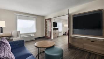 Habitación de hotel con sofá, una mesa redonda, televisor y puerta corredera hacia el baño.