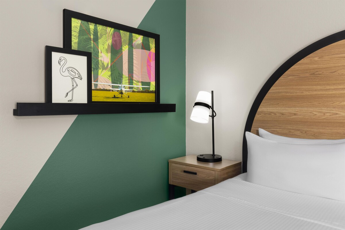 Quarto de hotel com cama, mesa de cabeceira com luminária, prateleira com obras de arte e uma parede verde de destaque.