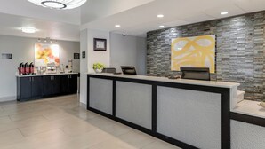 A reception area with a modern design, featuring a counter with a textured panel, a large abstract artwork on the wall, and a coffee station with various items. - Best Western Auburndale Inn & Suites (Auburndale)