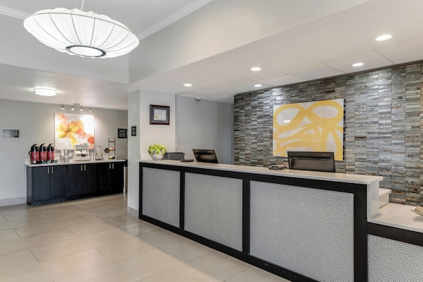 A reception area with a modern design, featuring a counter with a textured panel, a large abstract artwork on the wall, and a coffee station with various items.