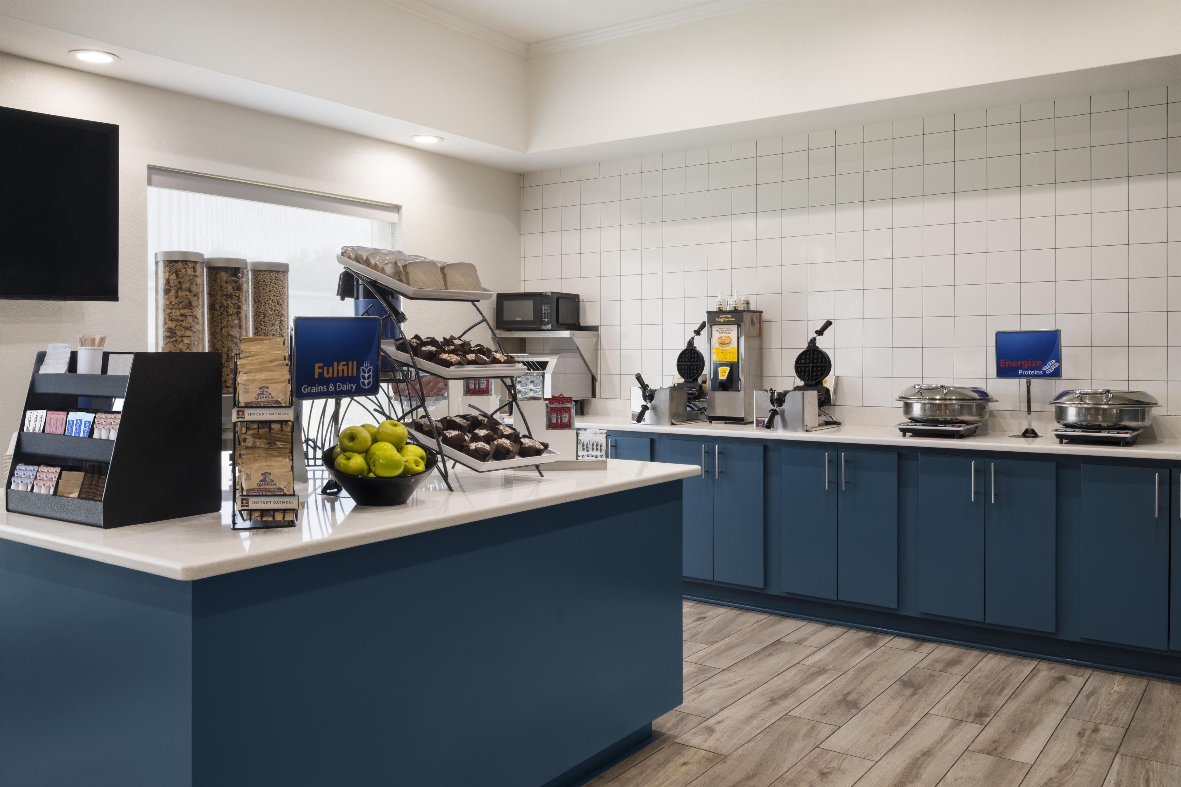 A self-service breakfast area with a variety of food options, including cereals, fruits, and a waffle maker.