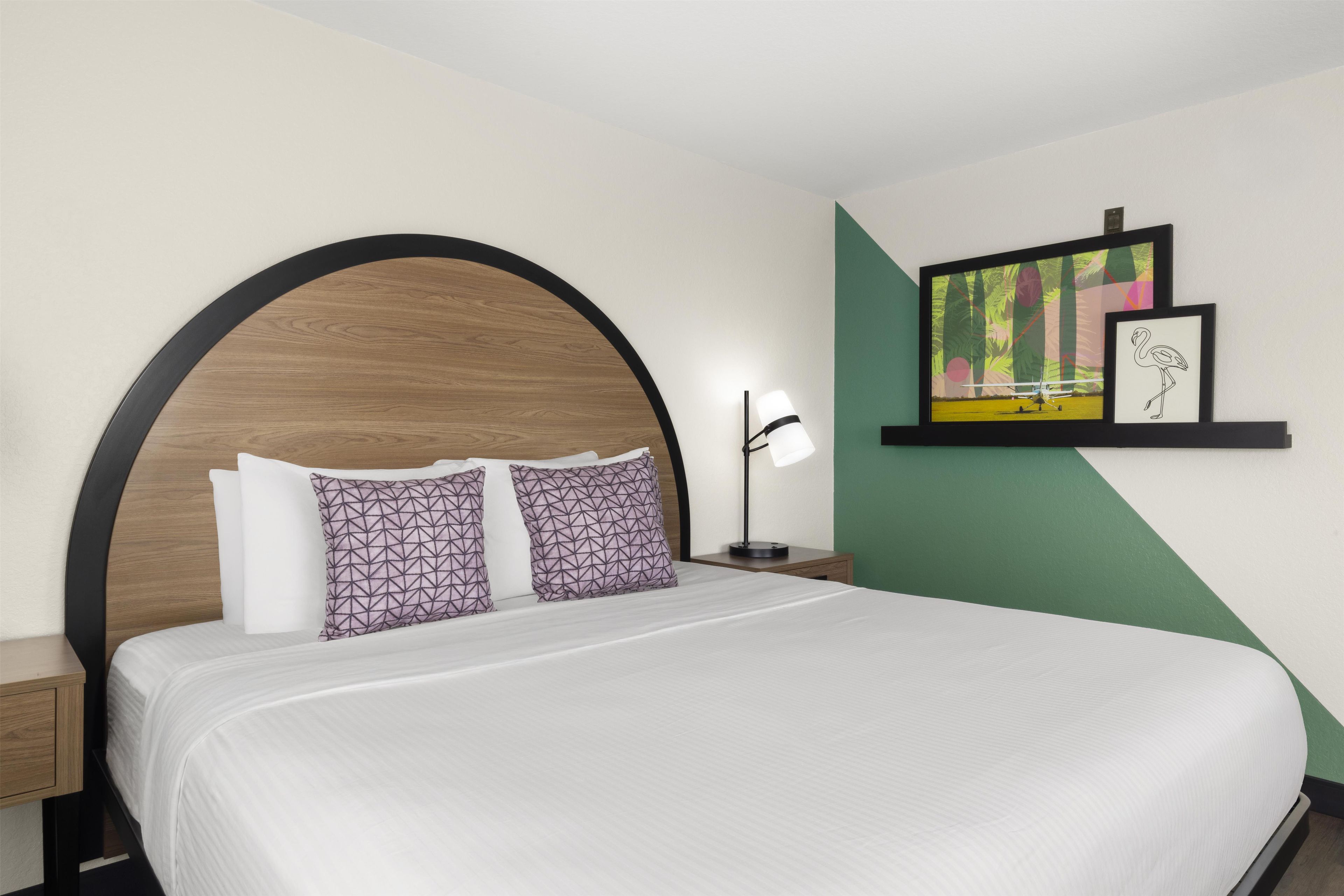 A hotel room with a large bed, two patterned pillows, a nightstand with a lamp, and a wall with artwork.
