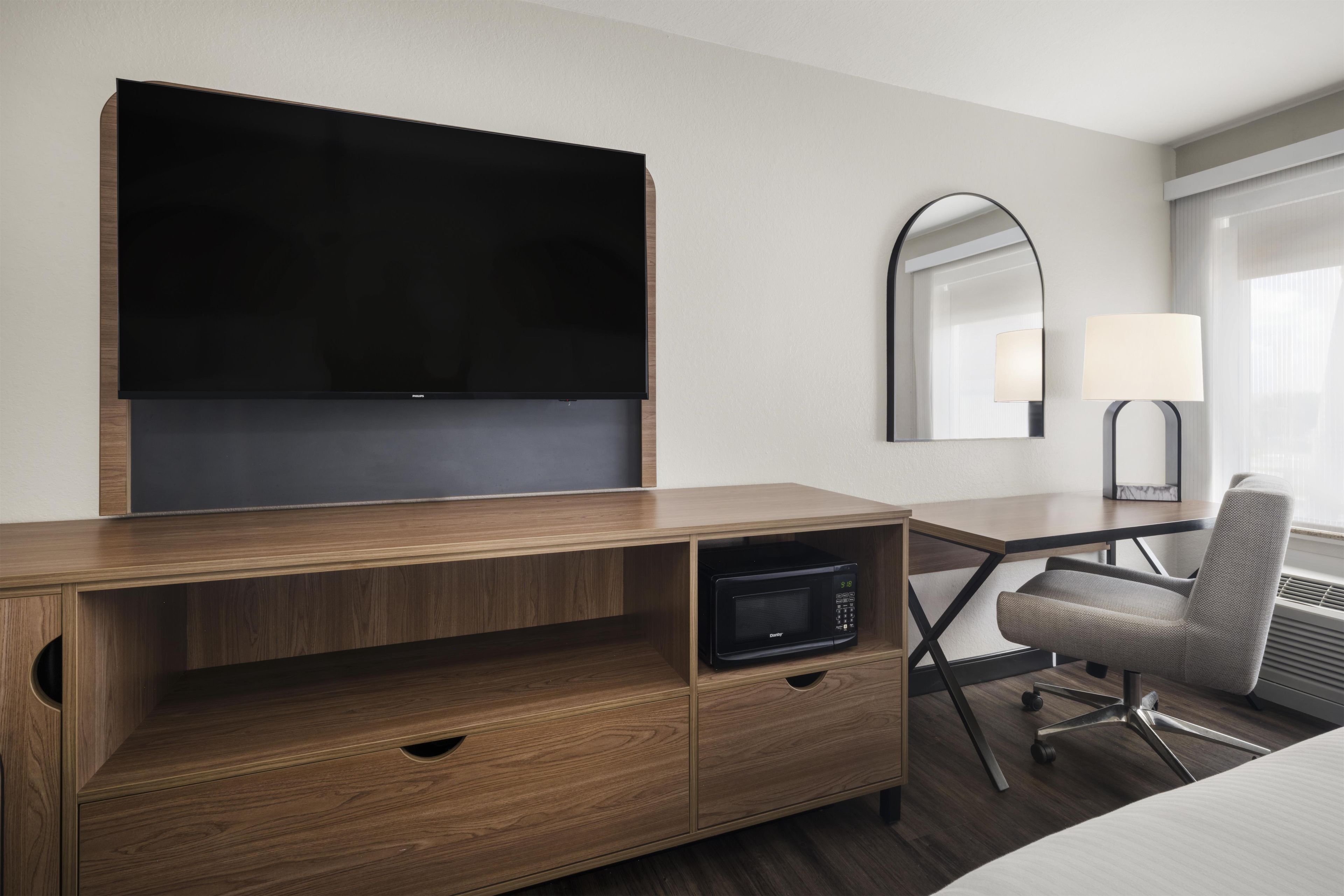 A hotel room with a wooden desk, a flat-screen TV, a microwave, a chair, a bedside table, and a lamp.