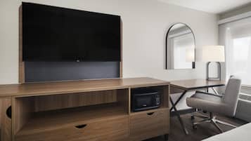 A hotel room with a wooden desk, a flat-screen TV, a microwave, a chair, a bedside table, and a lamp.