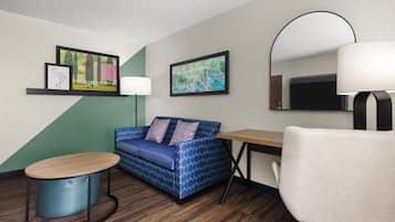 Quarto de hotel com sofá azul estampado, mesa de madeira redonda, poltrona branca, escrivaninha com abajur e duas molduras na parede.