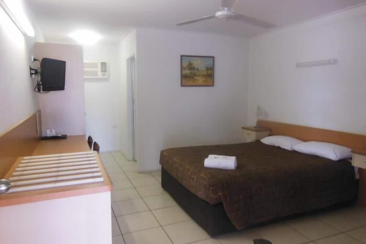 A hotel room with a bed, a desk, a chair, a ceiling fan, a painting, and a TV on the wall.