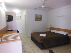A hotel room with a bed, a desk, a chair, a ceiling fan, a painting, and a TV on the wall. - Country Road Motel (Queenton)