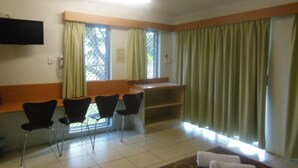 A hotel room with a ceiling fan, a flat-screen TV mounted on the wall, a desk with a chair, a window with curtains, and a seating area with chairs. - Country Road Motel (Queenton)