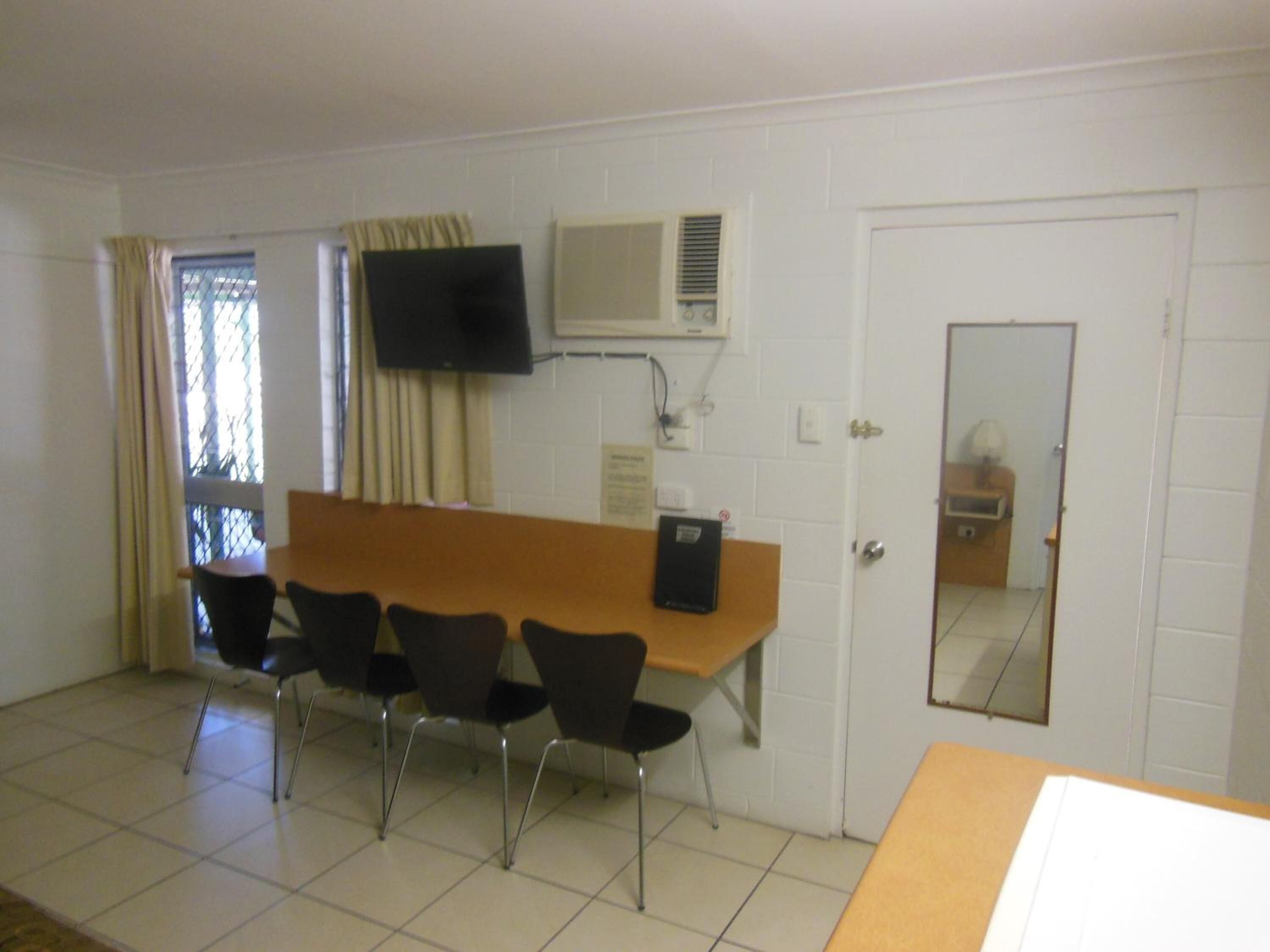Una habitación con una mesa larga, cuatro sillas, un televisor montado en la pared y una puerta con un espejo.