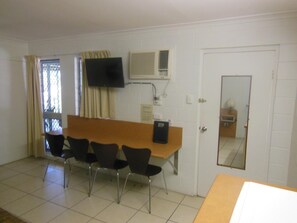 A room with a long table, four chairs, a TV mounted on the wall, and a door with a mirror. - Country Road Motel (Queenton)
