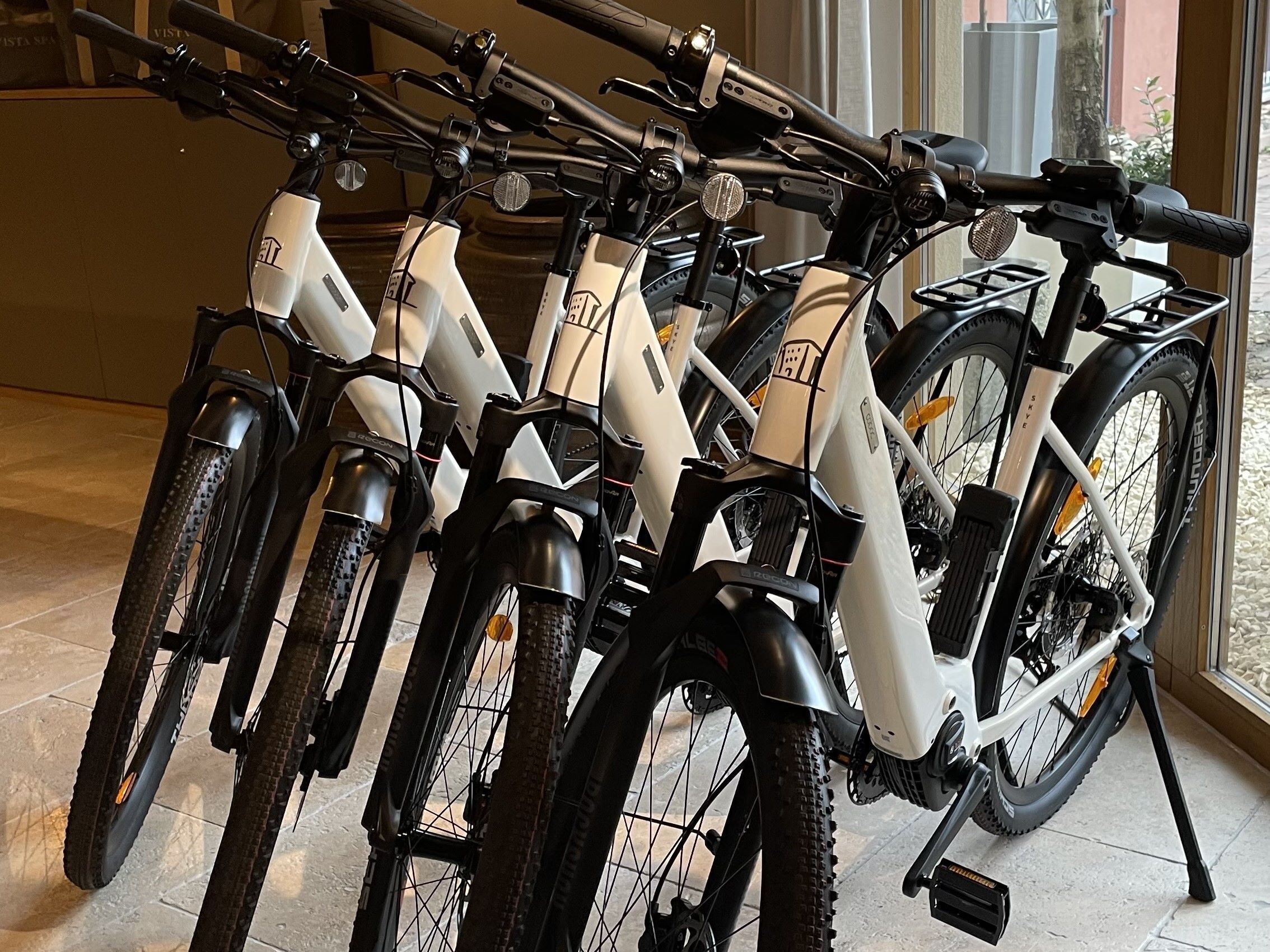 A row of electric bicycles parked indoors, with a tiled floor and a window in the background.