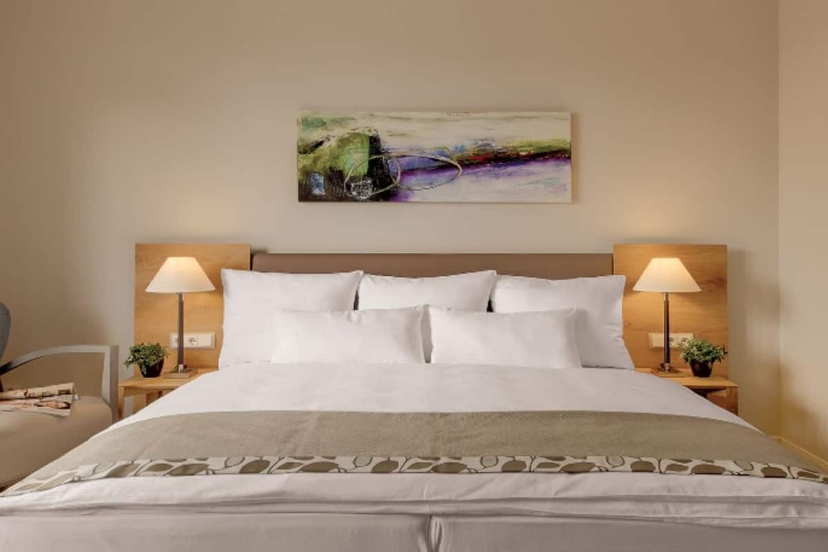 Une chambre d’hôtel avec un grand lit, deux lampes de chevet, une tête de lit, un tableau au-dessus du lit et une chaise avec un magazine.