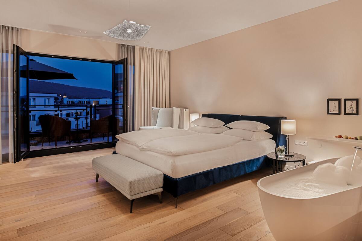 Une chambre d’hôtel moderne avec un grand lit, une baignoire et un balcon offrant une vue sur les immeubles.