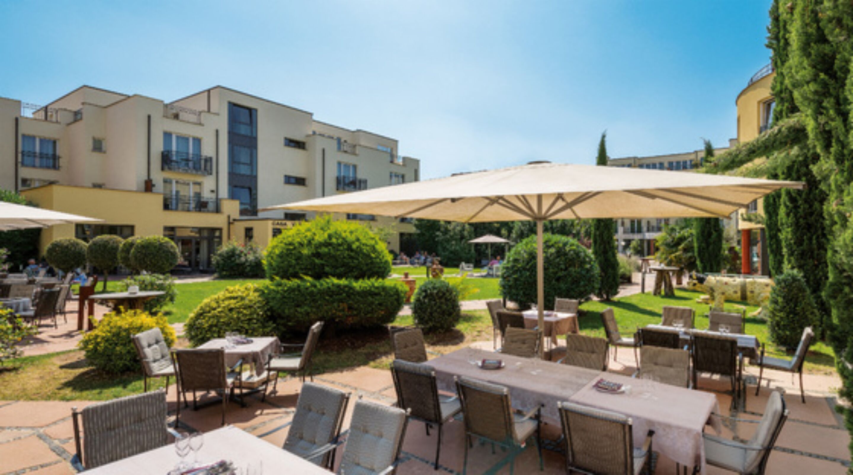 An outdoor patio area with tables and chairs, surrounded by greenery and a building in the background.