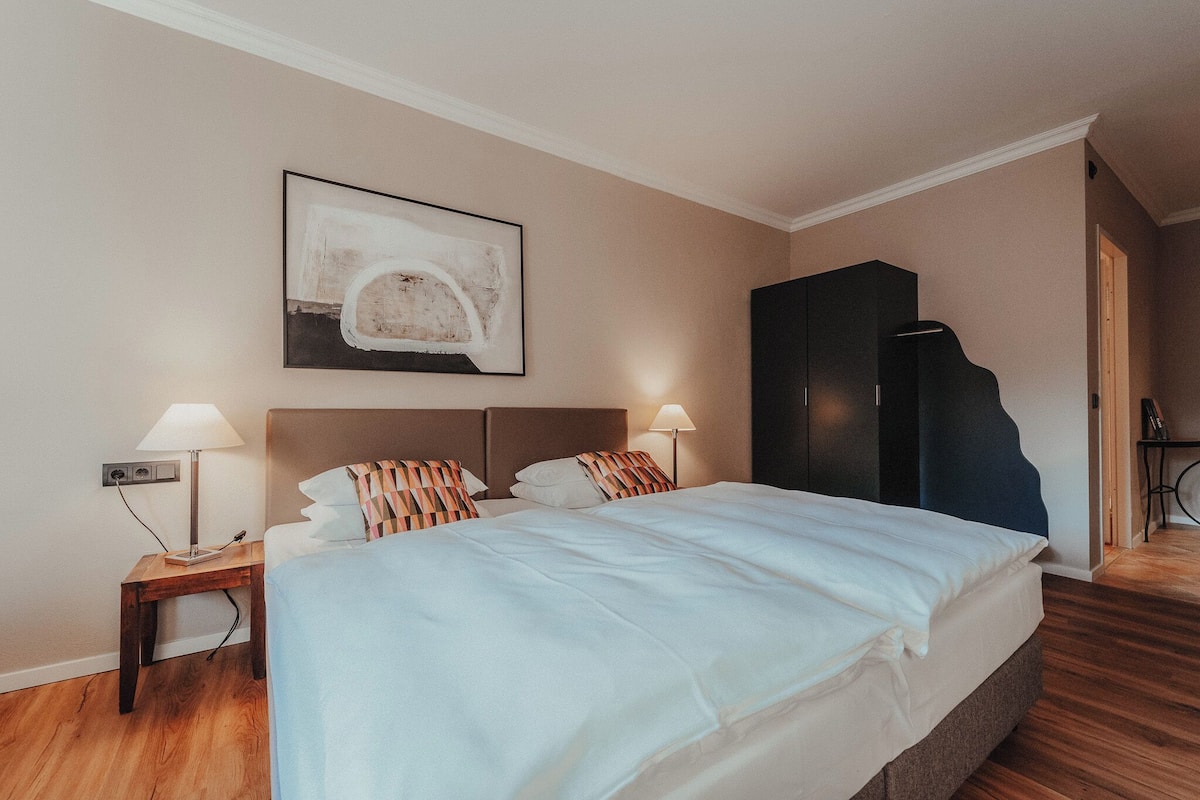 Une chambre d’hôtel avec un lit, deux lampes de chevet, un tableau abstrait accroché au mur, une armoire sombre et un plancher en bois.