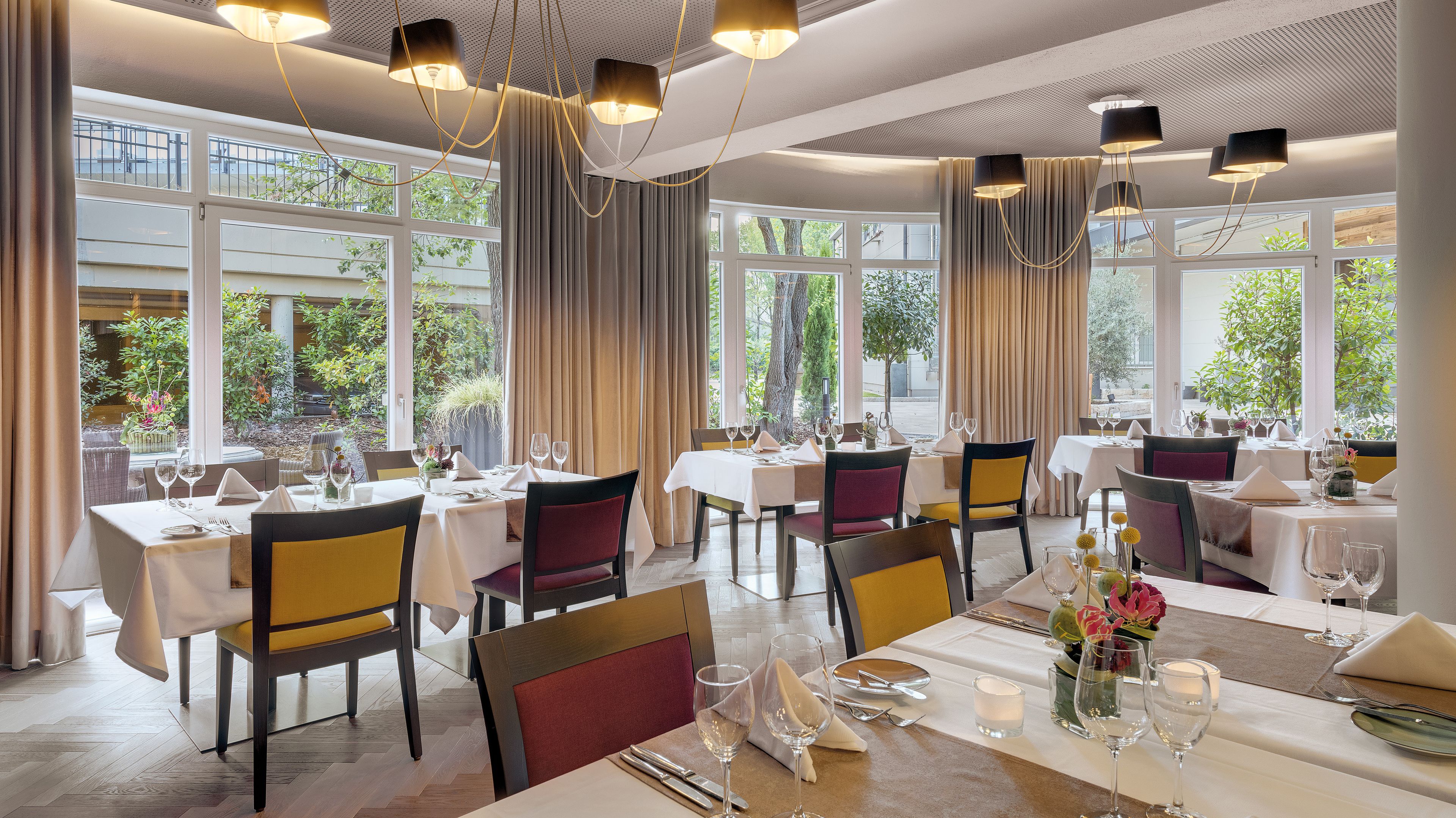 A restaurant with multiple tables set for dining, large windows offering views of greenery, and modern pendant lighting.