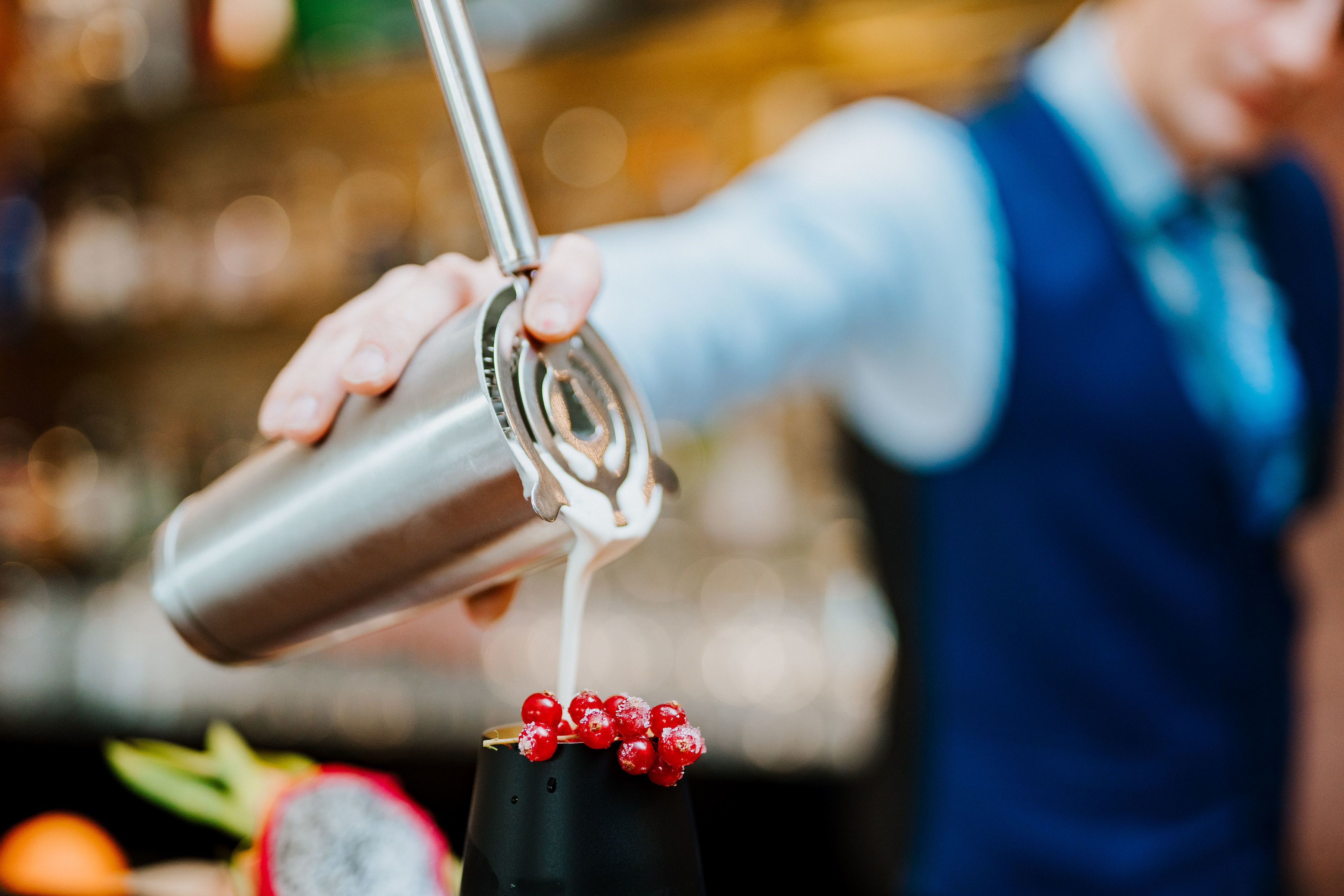 Un barman verse une boisson dans un verre dont le pied est noir et qui est décoré de baies rouges.