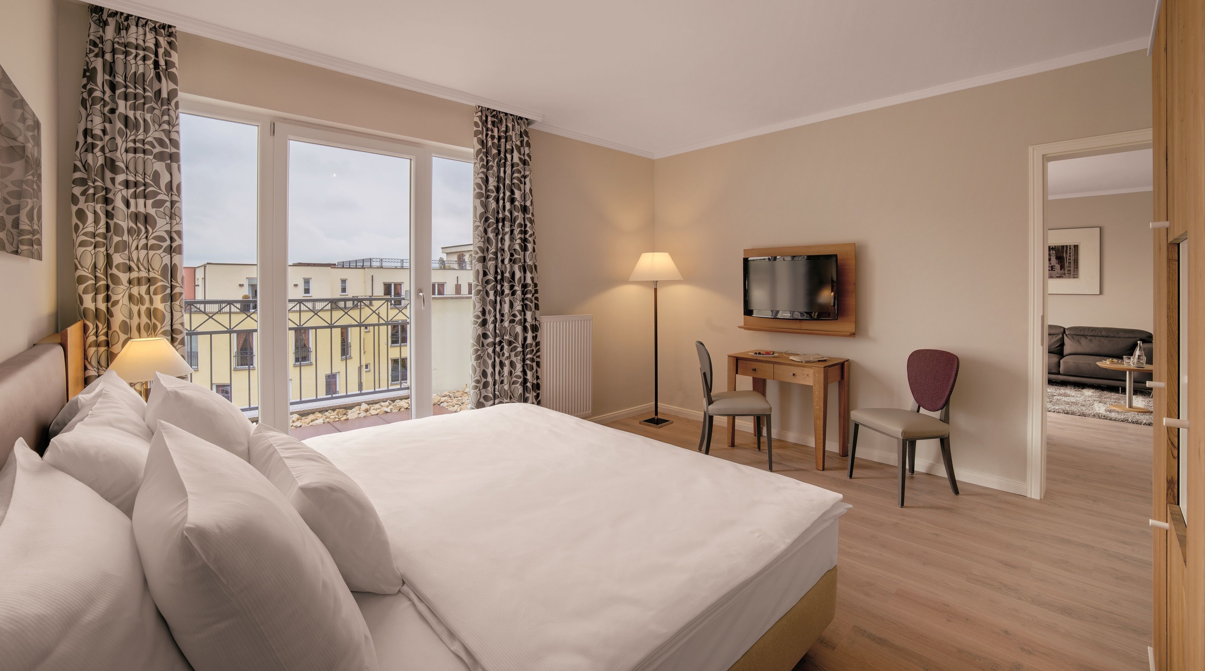 A hotel room with a bed, a desk, a chair, a TV, and a view of buildings through large windows.