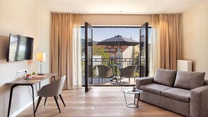 Ein modernes Hotelzimmer mit Balkon, einer Couch, einem Sessel, einem Schreibtisch und einem Fernseher.