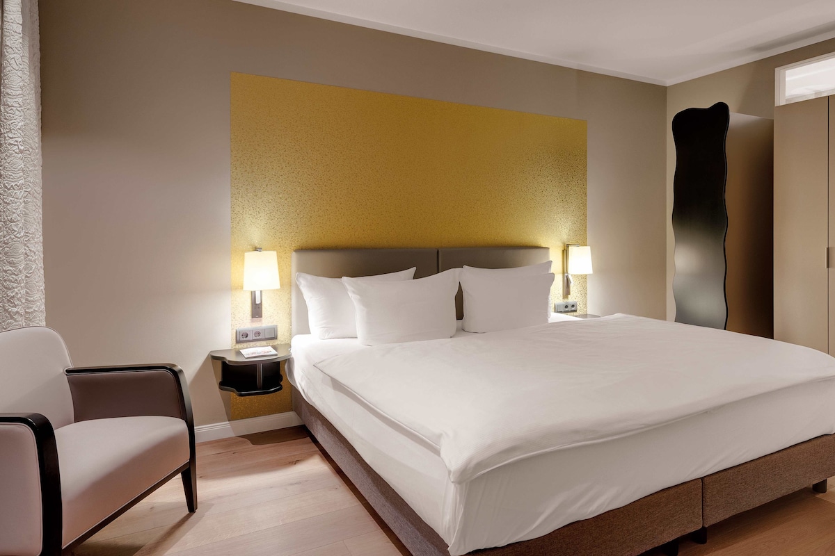 A hotel room with a large bed, two bedside tables with lamps, a chair, and a wardrobe.