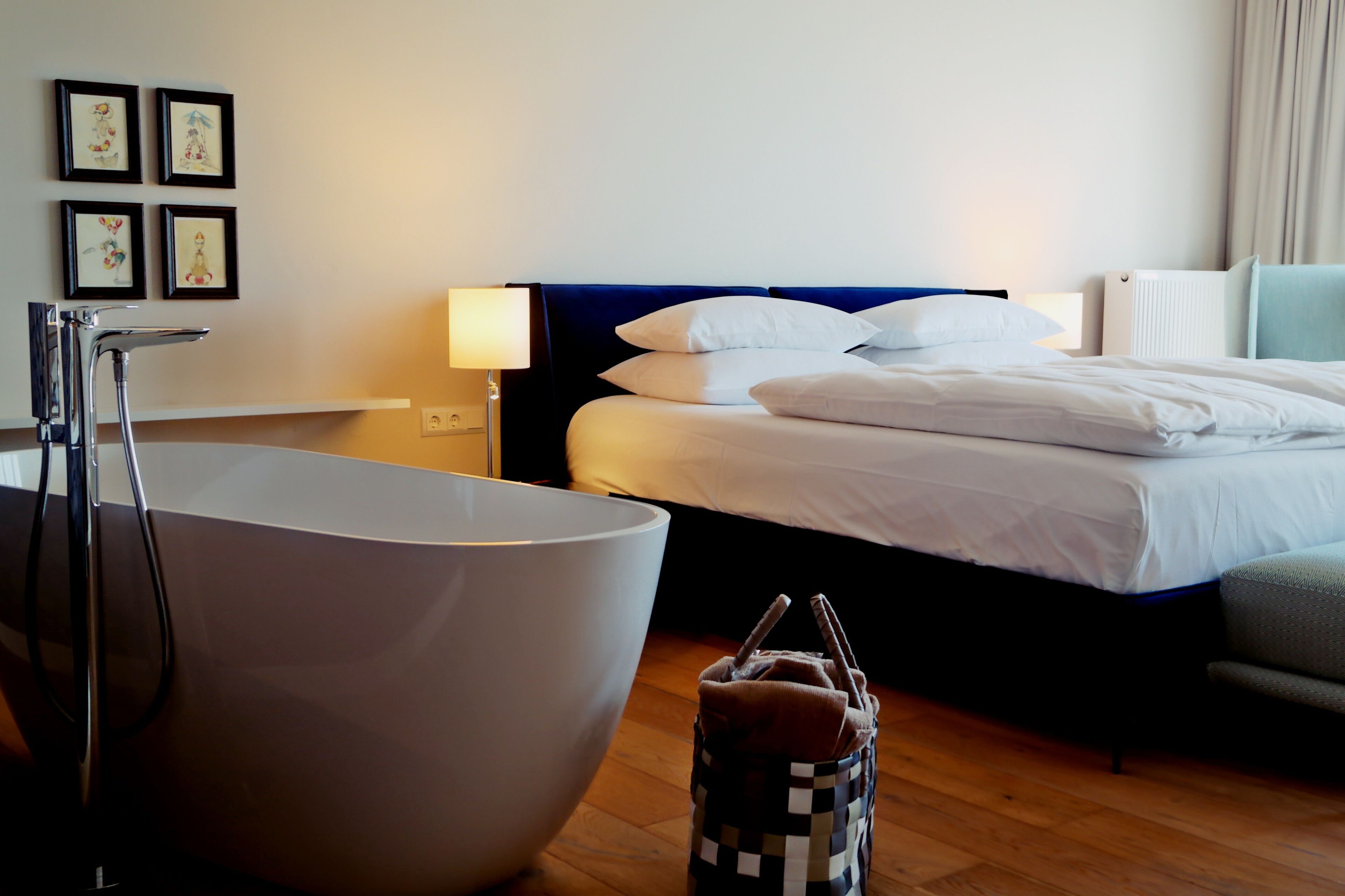 A hotel room with a large bathtub, a bed with white linens, a bedside table with a lamp, and a wall with framed artwork.