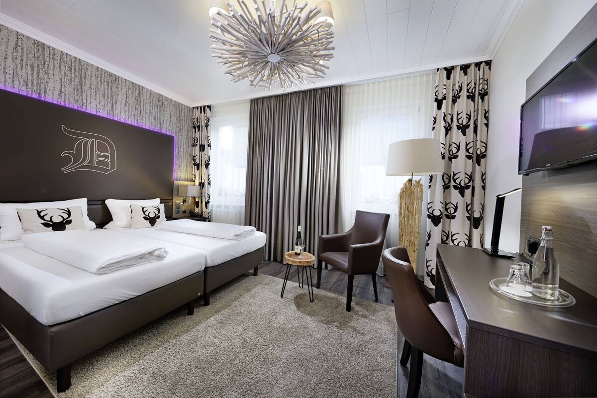 Ein Hotelzimmer mit einem großen Bett, einem Schreibtisch mit Fernseher, einem Sessel und einem kleinen Tisch mit Flasche und Gläsern.