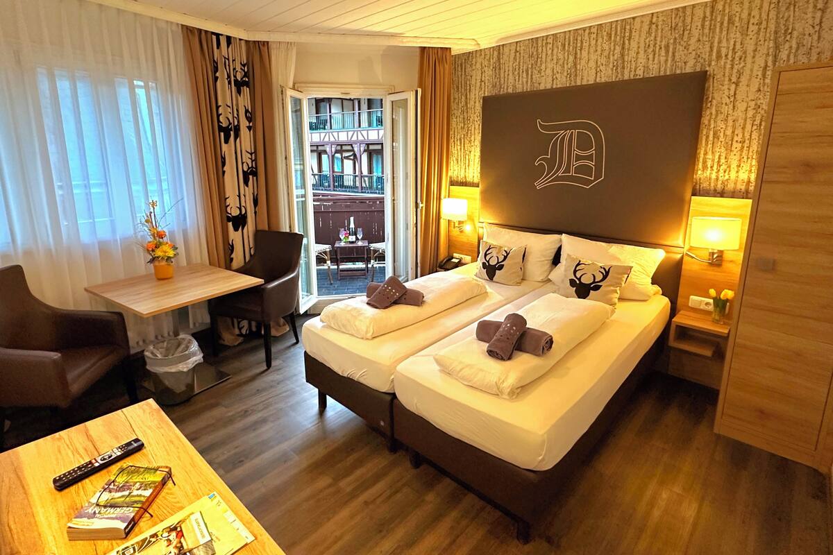 Ein Hotelzimmer mit Bett, zwei Nachttischen, einem Schreibtisch mit Stuhl, einem kleinen Tisch mit Fernbedienung und Büchern sowie einem Blick auf den Außenbalkon.