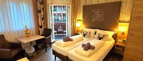 A hotel room with a bed, two bedside tables, a desk with a chair, a small table with a remote and books, and a view of the outside balcony.