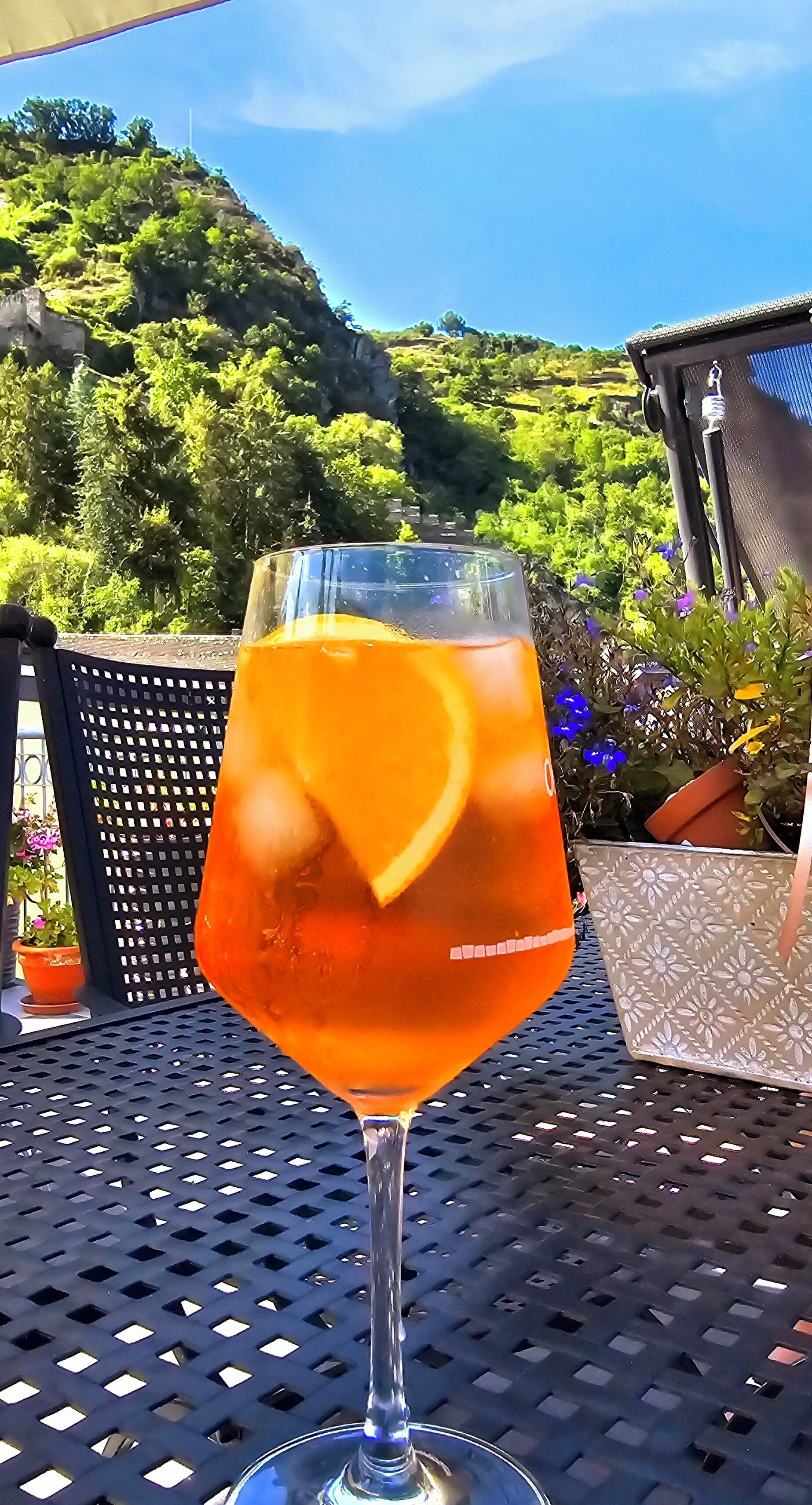 A glass of orange beverage with a slice of orange on a metal table outdoors.