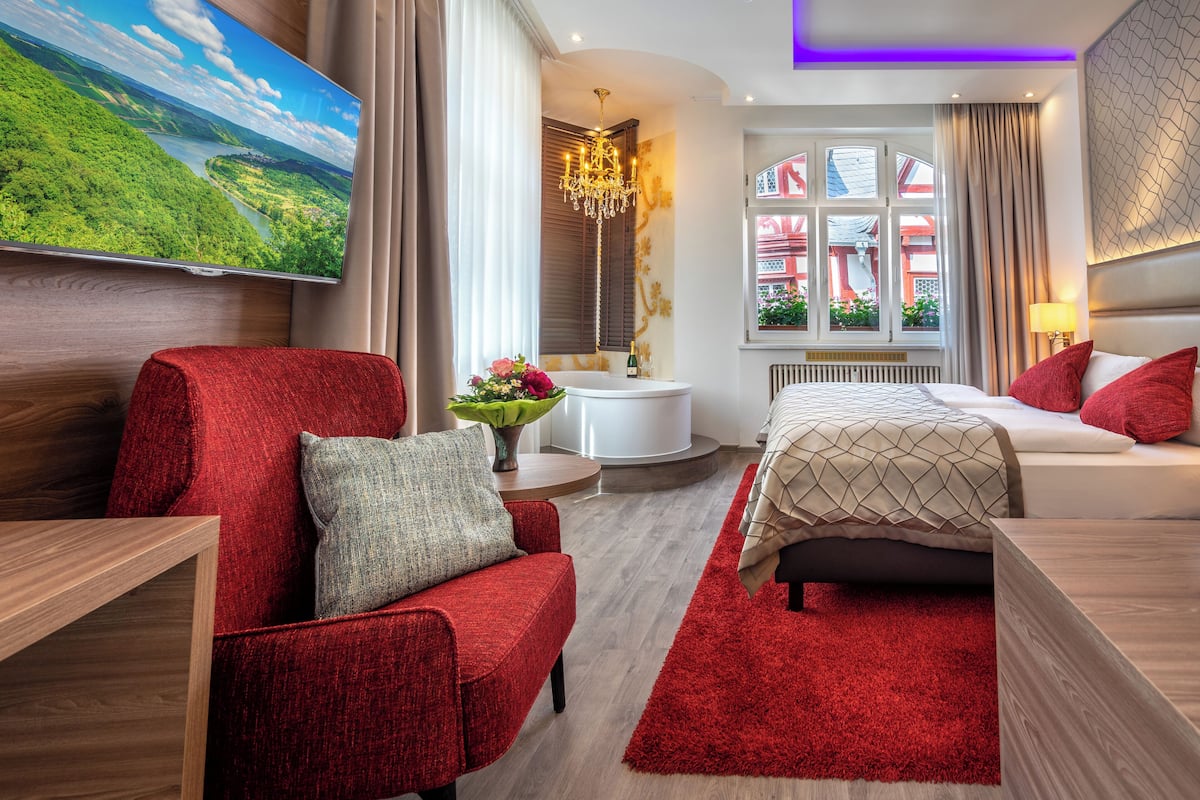 Ein modernes Hotelzimmer mit großem Fernseher, der eine schöne Landschaft zeigt, einem roten Sofa, einem Bett mit roten Kissen und einem kleinen runden Tisch mit Blumenvase.