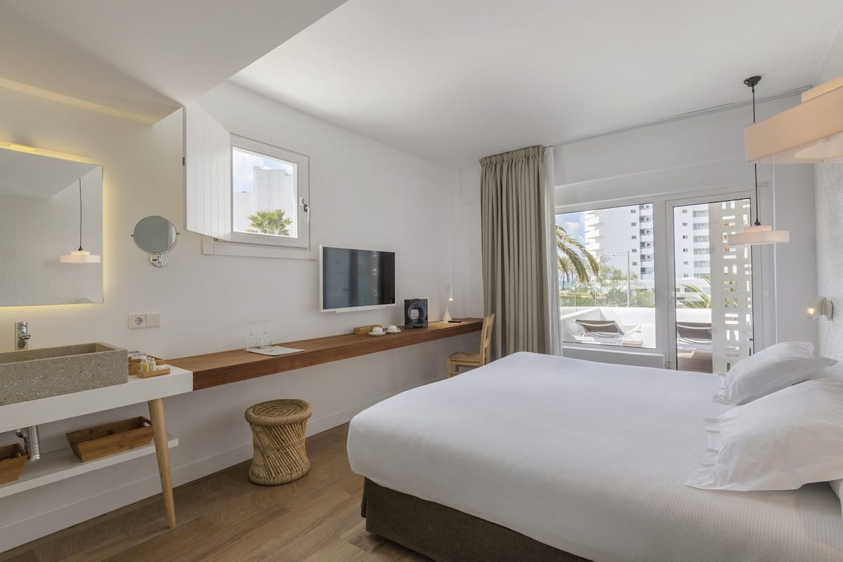 Habitación de hotel moderna con una cama grande, televisión integrada, tocador con lavamanos y balcón con vistas a palmeras.