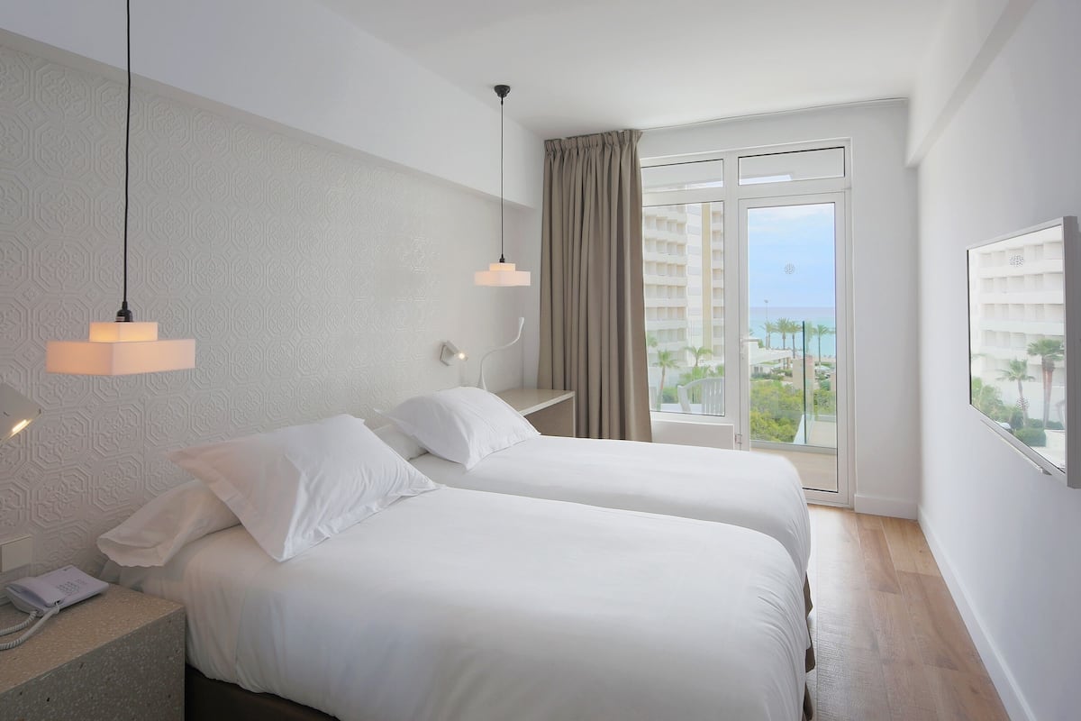 Habitación de hotel con dos camas, un teléfono en la mesita de noche, una ventana con vistas al mar y un papel tapiz estampado.