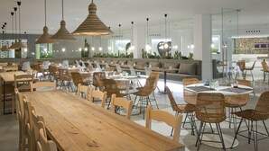 Un comedor amplio con varias mesas y sillas, iluminación colgante moderna y una zona de estar con sofá y cojines.