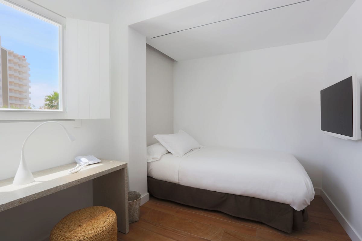 Un dormitorio con una cama, un escritorio con lámpara, una silla y una ventana con vista a edificios.