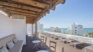 Een terras op het dak met een houten pergola, witte lounge fauteuils en tafels, met uitzicht op een strand en gebouwen.