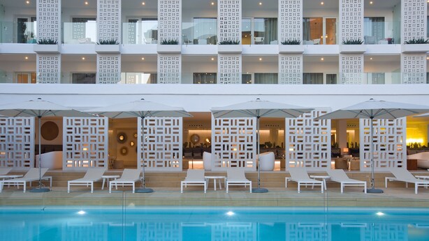 Un hotel con zona de piscina que cuenta con tumbonas y sombrillas, diseño arquitectónico moderno con balcones blancos y una piscina azul cristalina.