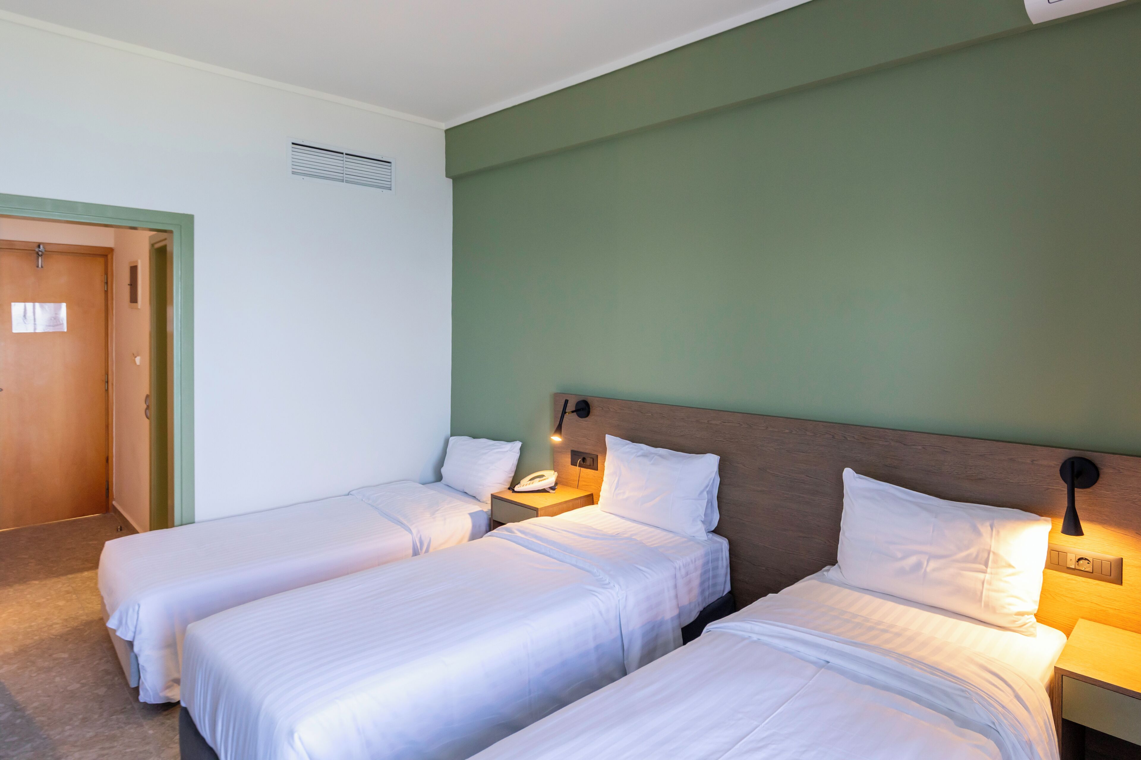 A hotel room with two single beds, a wooden headboard, a nightstand with a lamp, and a green accent wall.