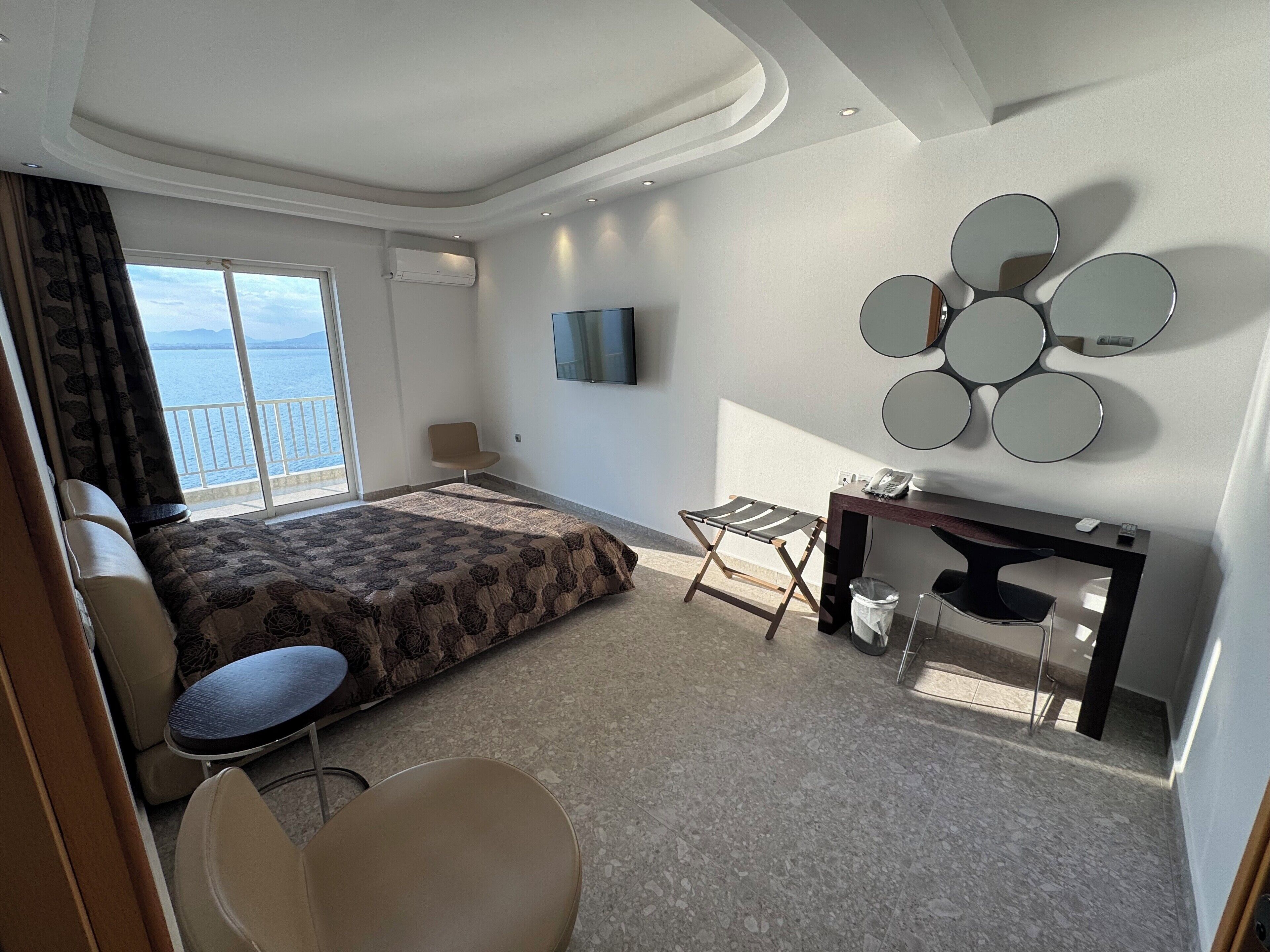 A hotel room with a bed, a desk, a chair, a TV, and a view of the sea.