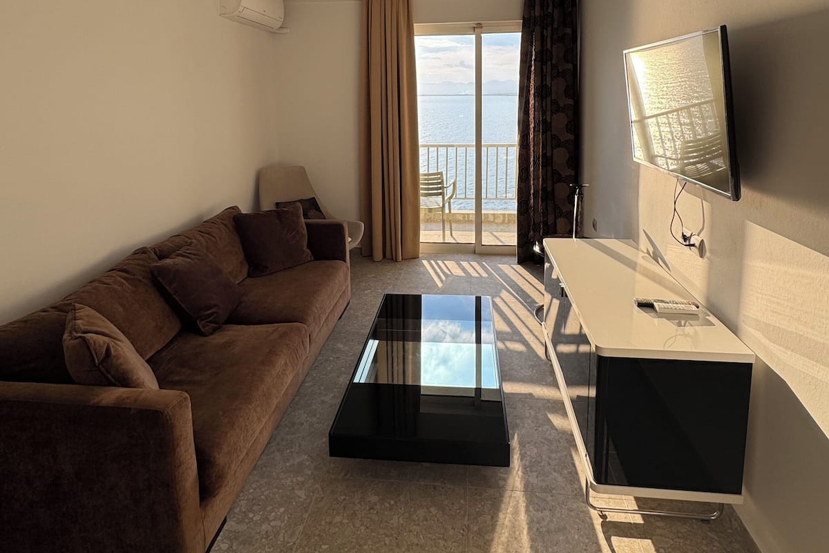Habitación de hotel con sofá, mesa de centro de vidrio, televisión y balcón con vista al mar.