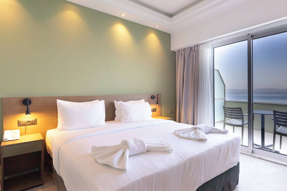 Habitación de hotel con una cama grande, una mesita de noche con teléfono y vistas al mar desde un balcón.