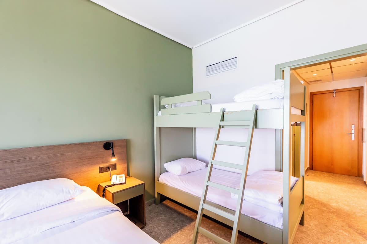 Habitación con literas, una escalera de madera, una mesita de noche con lámpara y una puerta a otra habitación.