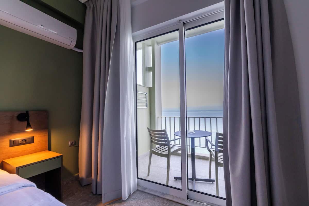 Habitación de hotel con cama, mesita de noche, escritorio, silla, mesa y balcón con vista al mar.