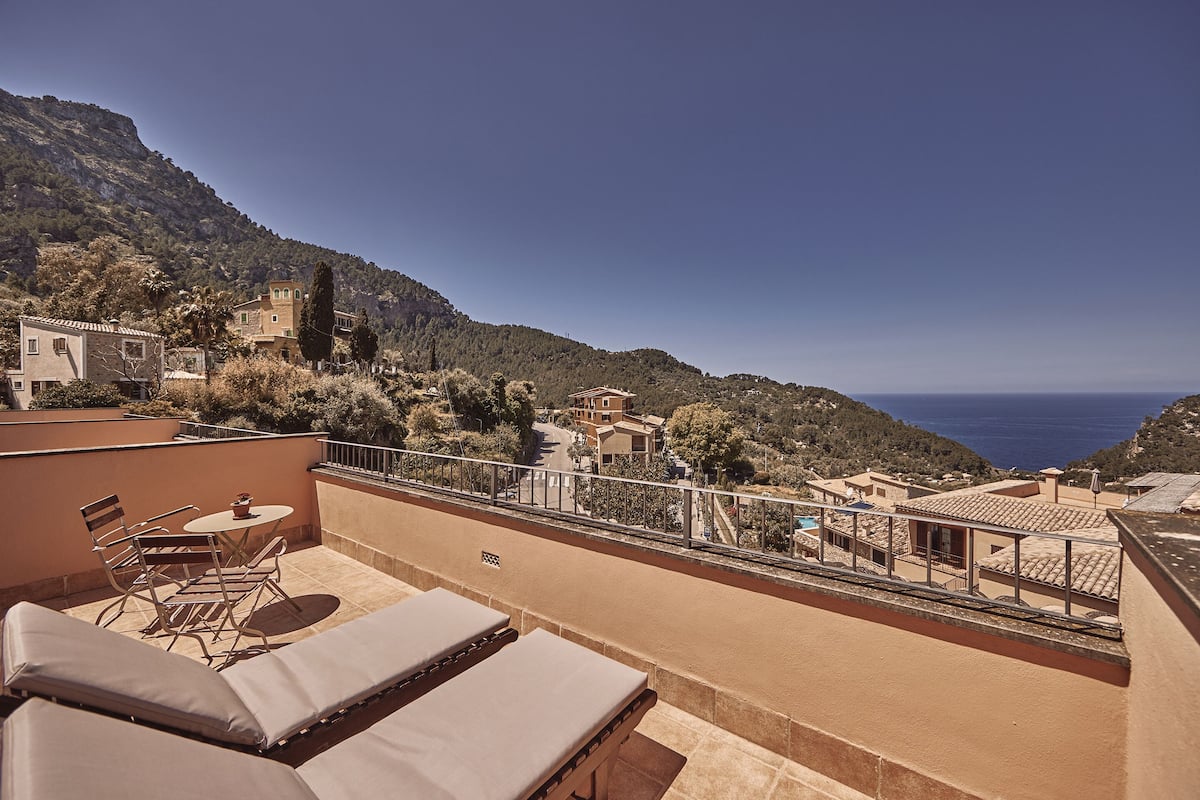 A rooftop terrace with lounge chairs and a table, overlooking a coastal village and the sea.
