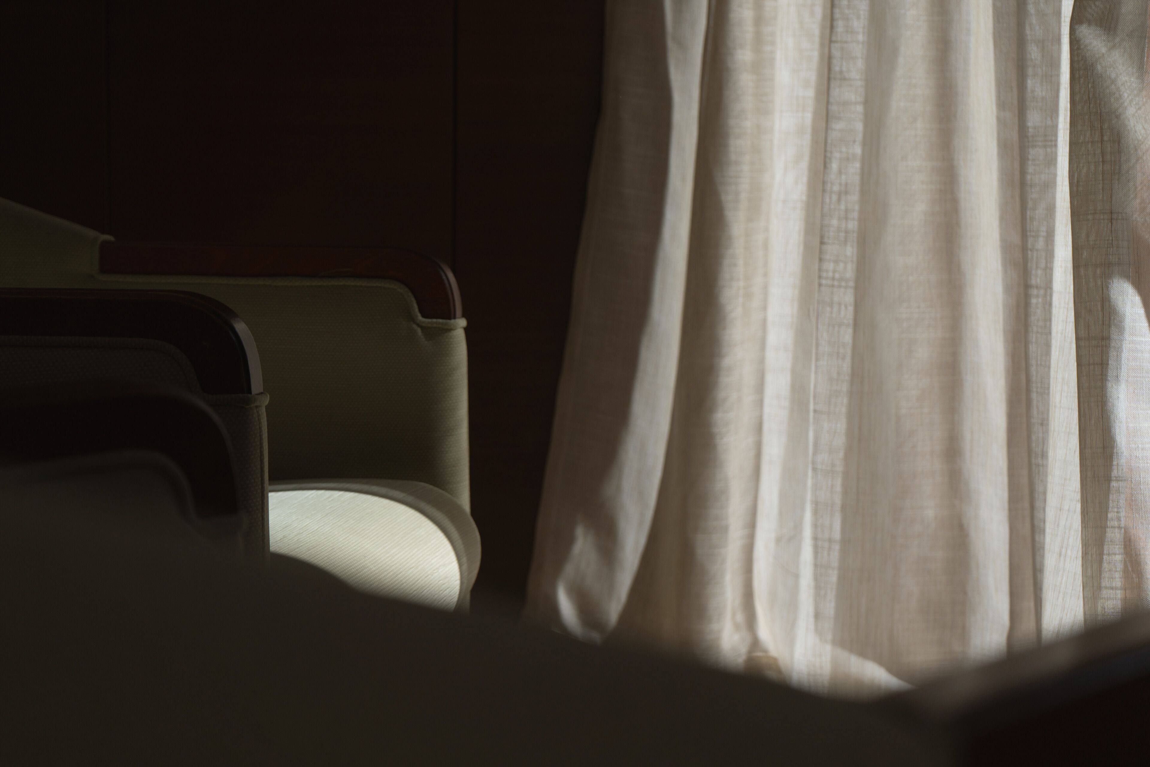 Une pièce faiblement éclairée, avec une chaise et un rideau à moitié tiré, laissant filtrer un peu de lumière.