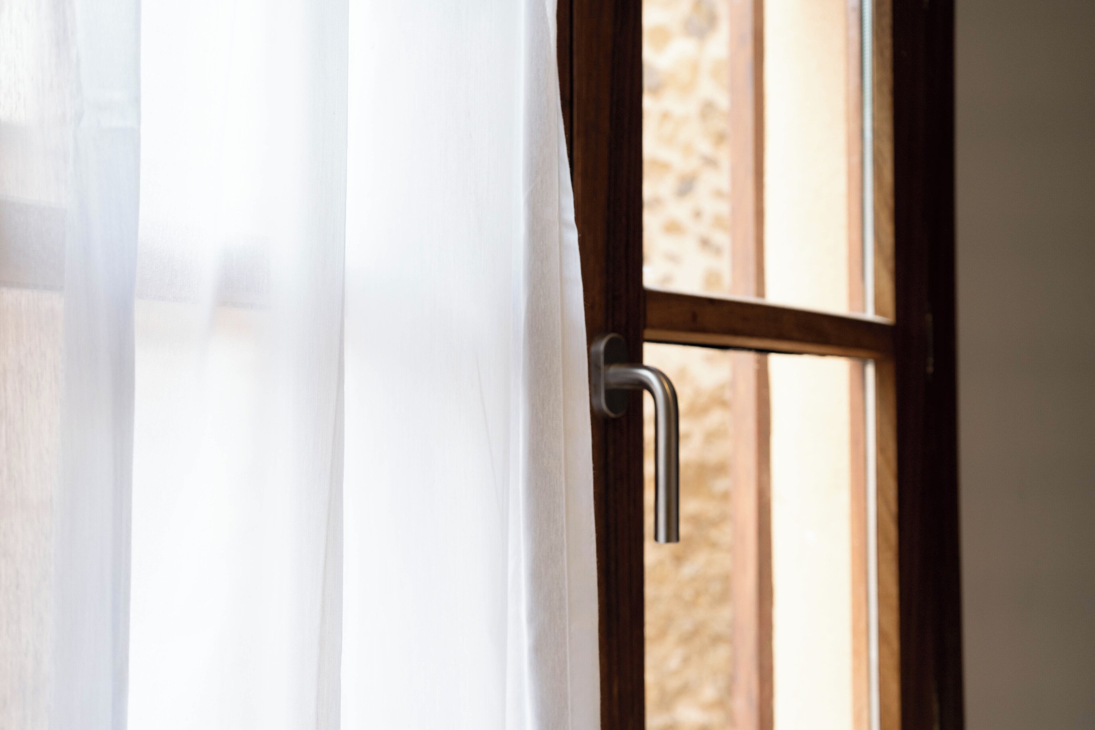 A window with a metal handle and a wooden frame, partially covered by a sheer curtain.