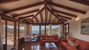 Un vaste espace de vie comprenant un grand canapé, une table basse et une table à manger avec des chaises. De grandes fenêtres offrent une vue sur la mer et les collines.