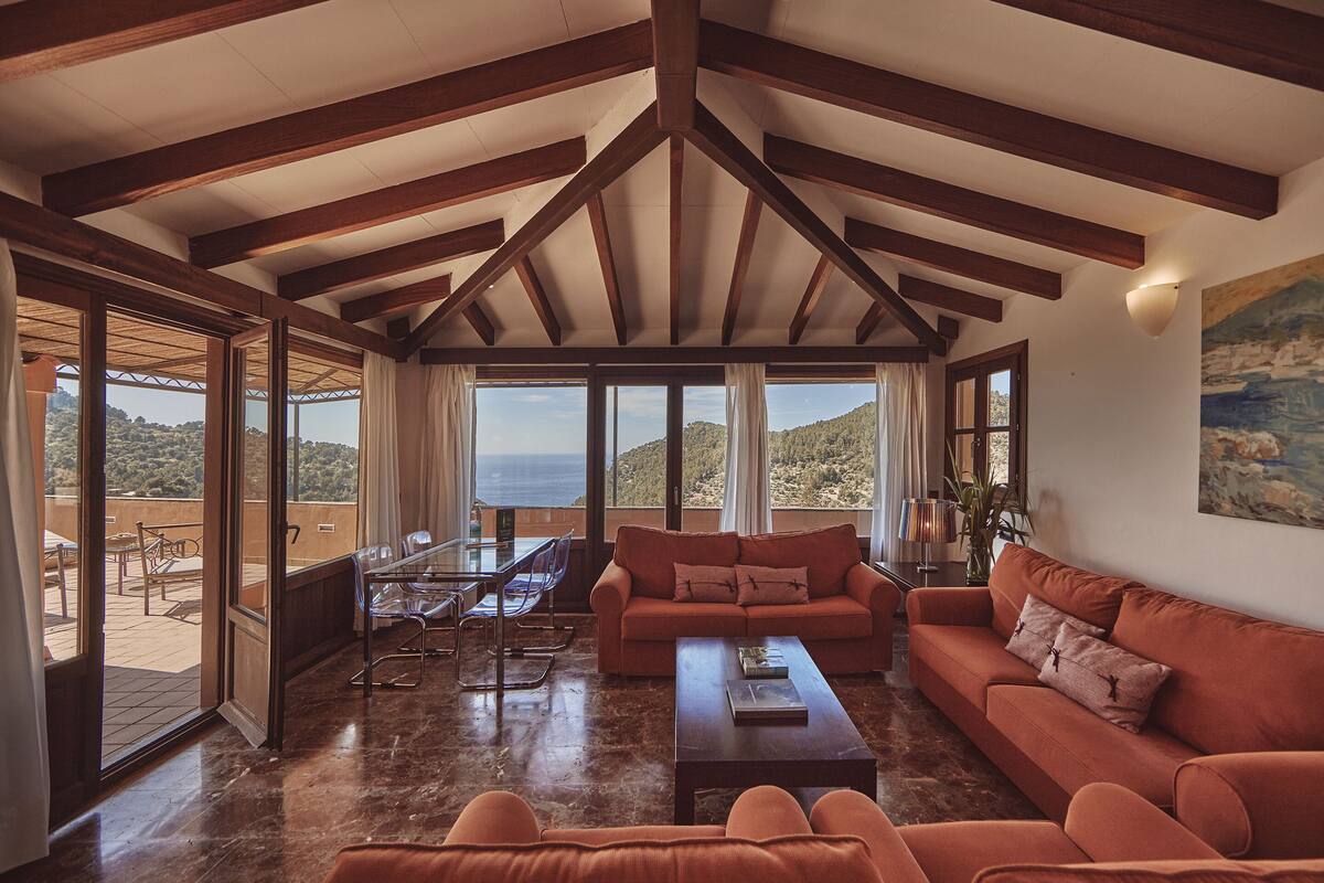 A spacious living area with a large sofa, a coffee table, and a dining table with chairs. There are large windows offering a view of the sea and hills.