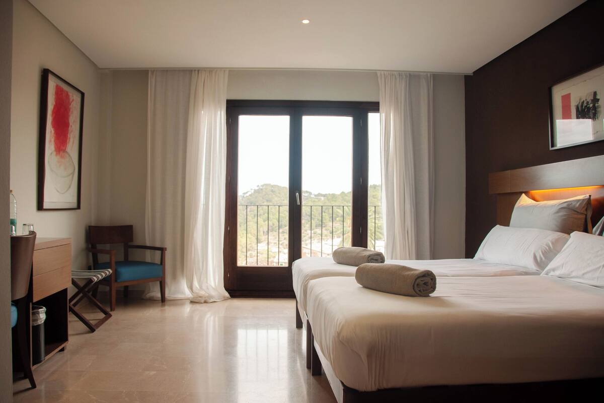 A hotel room with a large bed, a chair, a desk, and a view of the outdoors through a large window.