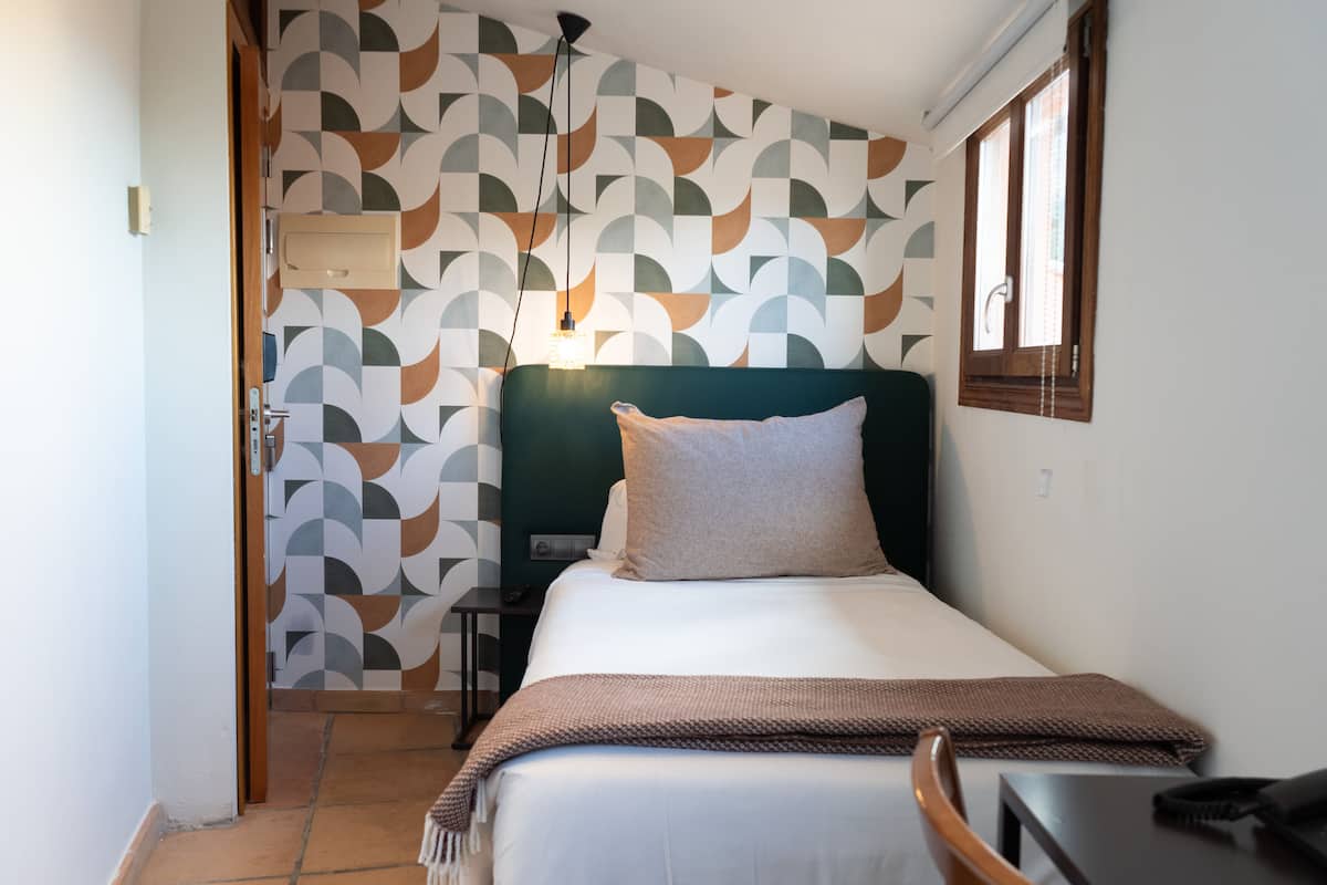 A hotel room with a patterned wall, a bed with a green headboard, a bedside table, and a window.